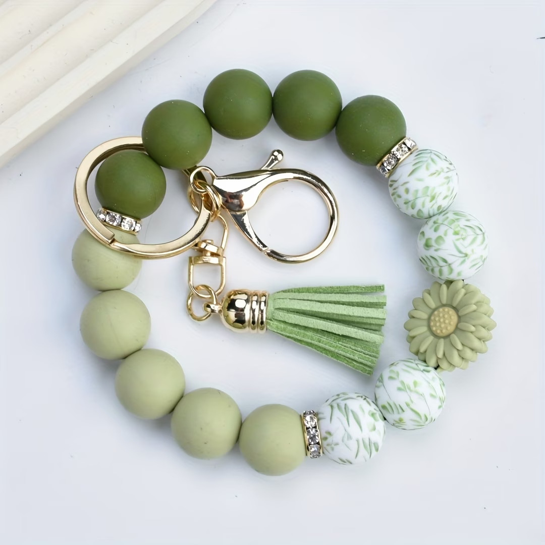 A handcrafted beaded wristlet keychain from Woodland Beads & Co featuring a gradient of olive and sage green beads, white beads with green leaf prints, a sage green sunflower charm, and a matching green tassel with gold hardware.