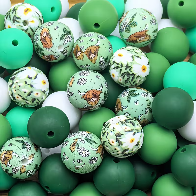 A collection of premium silicone beads featuring hand-drawn Highland Cow prints on sage green, white daisy botanical patterns, and a mix of solid forest green, mint, and white beads by Woodland Beads & Co.