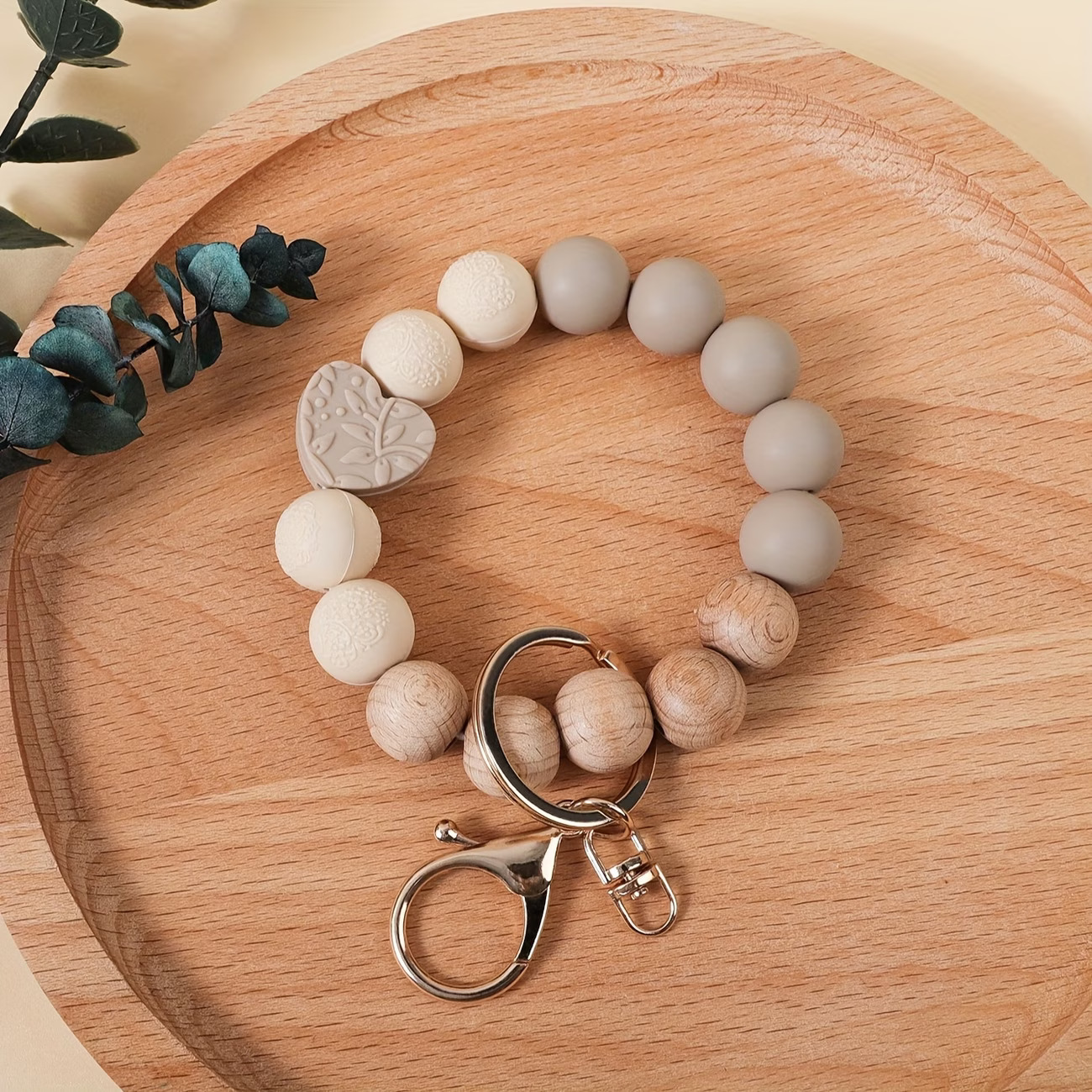 Hand-created beaded wristlet keychain by Woodland Beads & Co featuring matte taupe and cream beads, natural timber-style beads, and a taupe heart-shaped focal bead with an embossed leaf pattern, shown on a wooden tray.