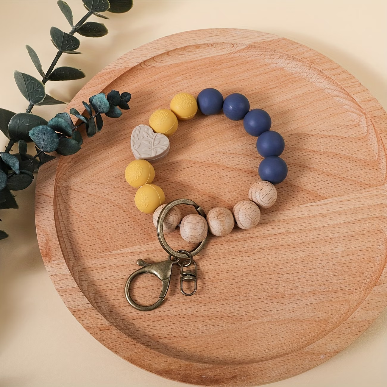 Hand-created beaded wristlet keychain by Woodland Beads & Co featuring matte navy and mustard yellow beads, natural timber-style beads, and a taupe heart focal bead with a leaf pattern, displayed on a wooden tray.