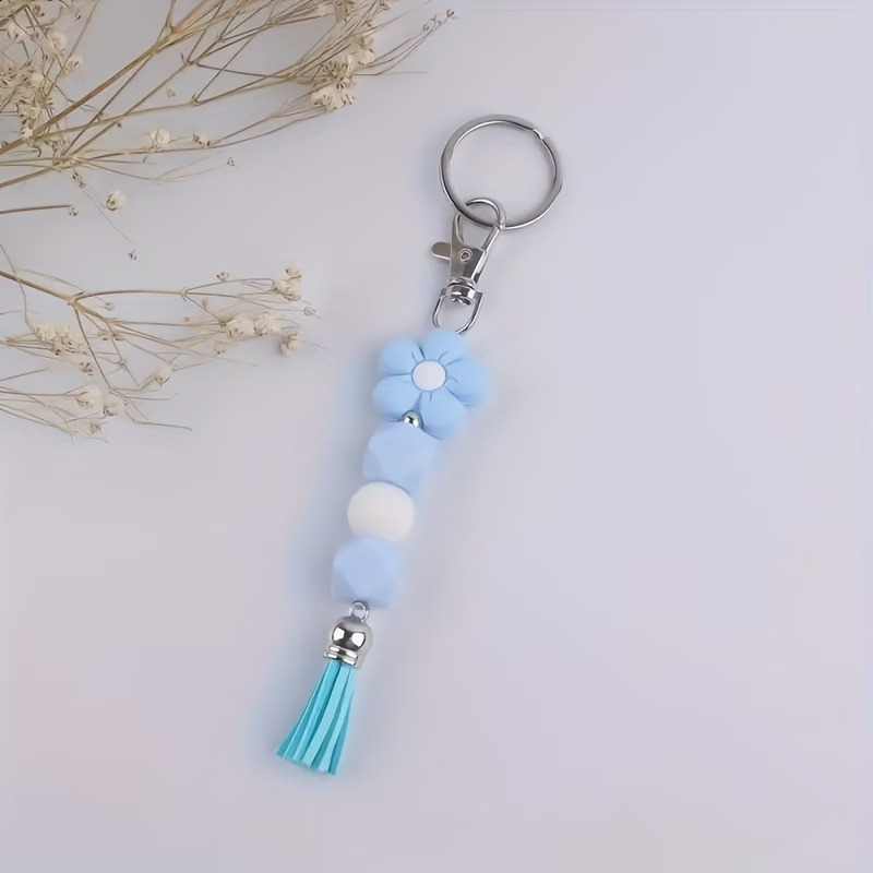 A blue and white beaded keychain featuring a silicone flower charm and a blue tassel. The beads include two light blue hexagons and one white sphere. It is attached to a silver swivel clasp and key ring, shown on a white background with dried floral accents.