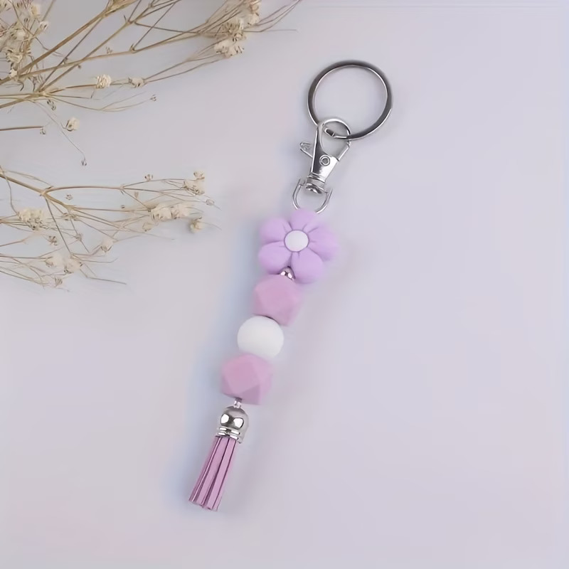 A lilac and white beaded keychain featuring a soft-touch silicone flower charm and a purple tassel. The design includes two lilac hexagonal beads and one white round bead on a silver connector. It is attached to a silver-tone swivel lobster clasp and key ring, displayed on a white surface with dried floral accents.